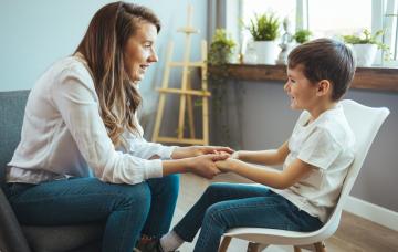 Terapia escolar para prevenir el TDAH en niños Mujer psicóloga coge de las manos a un niño sentado en una silla que sonríe