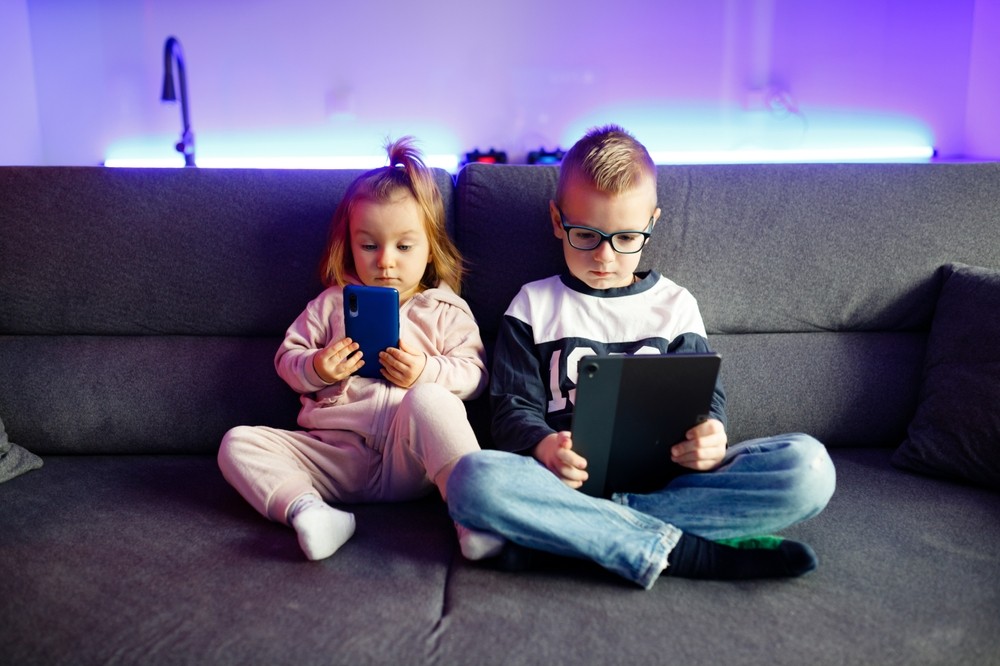 Abuso de pantallas en menores de 6 años Hermanos pequeños en el sofá viendo una tablet y un smartphone