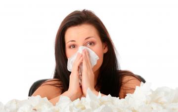 Qué es la rinitis Una mujer con rinitis se suena la nariz junto a una pila de pañuelos de papel usados