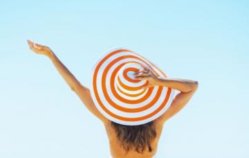 Cómo proteger tu cuero cabelludo y tu pelo de los rayos solares Chica de espaldas se sujeta el sombrero en la playa