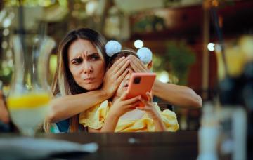 Pantallas de teléfonos negativas para los niños Madre tapando los ojos de su hija pequeña mientras esta mira un teléfono móvil