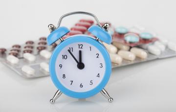 Hora a la que tomar la medicación Reloj azul marca casi las 12 y detrás varios blíster de medicamentos