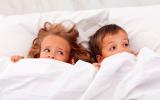 Niña y niño asustados en la cama tapándose la boca con la sábana