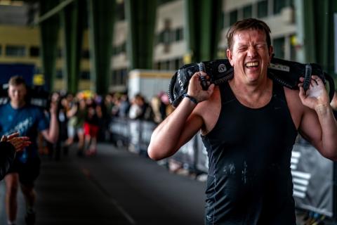 Hyrox: hombre con gestos de esfuerzo compite en Berlín Hombre carga con un pesado saco en una carrera de hyrox