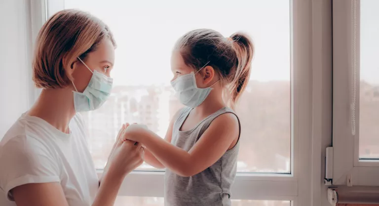 Madre e hija con mascarilla junto a la ventana