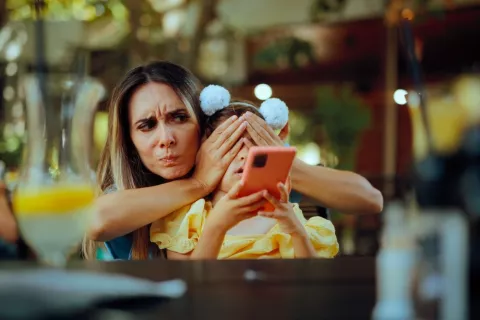 Pantallas de teléfonos negativas para los niños Madre tapando los ojos de su hija pequeña mientras esta mira un teléfono móvil