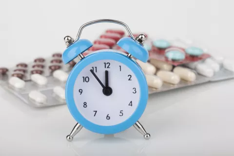 Hora a la que tomar la medicación Reloj azul marca casi las 12 y detrás varios blíster de medicamentos
