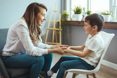Terapia escolar para prevenir el TDAH en niños Mujer psicóloga coge de las manos a un niño sentado en una silla que sonríe