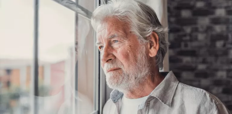 Hombre mayor con barba y canas mirando preocupado por la ventana