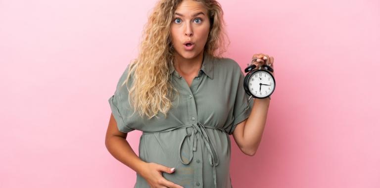 Mujer rubia embarazada con cara de sorpresa y un reloj en la mano