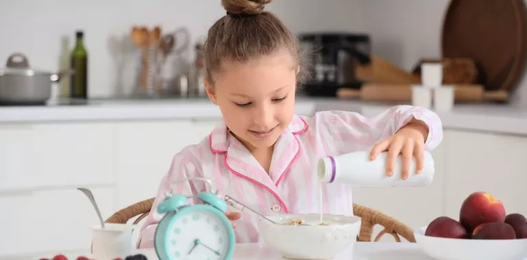 Niña en pijama se sirve leche en los cereales con un reloj que marca las 7 de la mañana