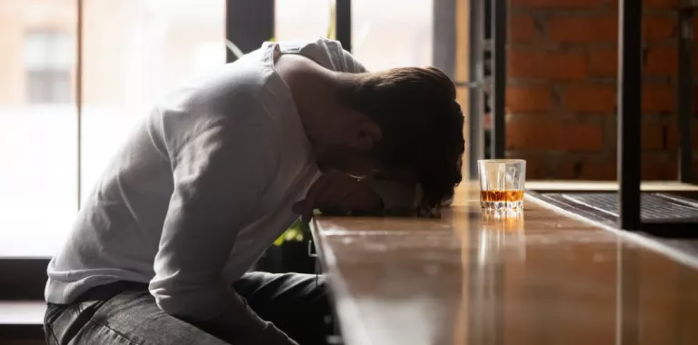 Joven ebrio apoya su cabeza sobre la barra del bar junto a un vaso de wisky