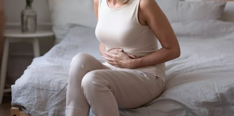 Mujer de mediana en la edad sentada en la cama con las manos en el abdomen dolorido