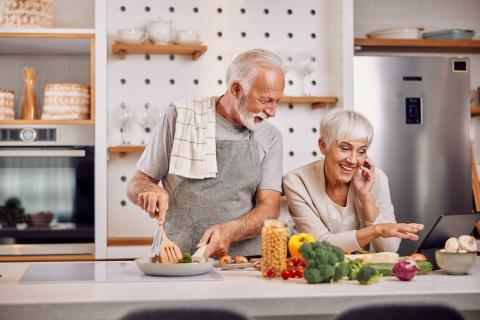 Pareja de adultos mayores cocinan alimentos saludables consultando una tablet
