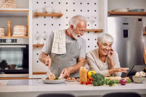 Pareja de adultos mayores cocinan alimentos saludables consultando una tablet