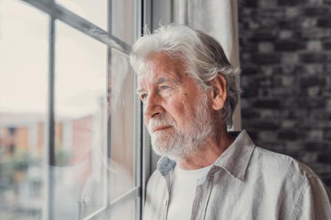 Hombre mayor con barba y canas mirando preocupado por la ventana