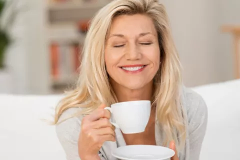 Mujer sonríe mientras sostiene una taza de café Mujer de mediana edad sonríe mientras se dispone a tomarse un café sentada en su sofá