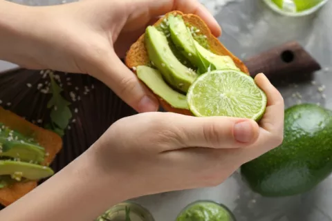 Mujer sostiene una tostada de pan integral con trozos de aguacate Manos de mujer preparando. una tostada con aguacate y lima