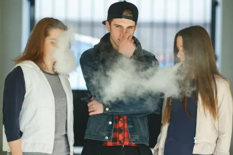 Dos chicas adolescentes vapean alrededor de un joven que se tapa la nariz