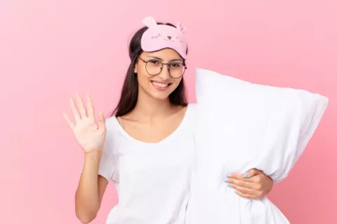 Chica con un antifaz en la frente y sujetando una almohada se despide sonriente antes de irse a la cama