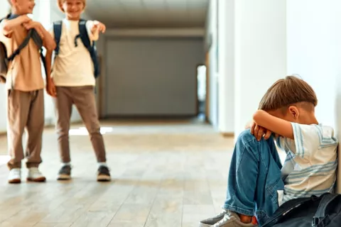 Niño sentado en el suelo de la escuela acosado por dos compañeros Niño sentado en el suelo se tapa la cara mientras dos compañeros de escuela se ríen de él