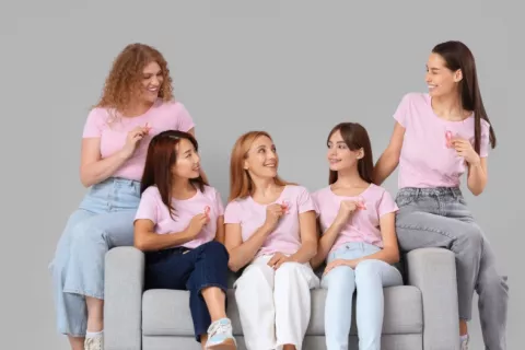Cinco mujeres jóvenes con camisetas y cintas rosa que simbolizan la lucha contra el cáncer de mama Grupo de mujeres jóvenes con cintas rosadas contra el cáncer de mama sentadas en un sofá