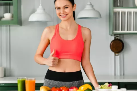 Una joven con ropa deportiva se dispone a desayunar zumos y frutas variadas Mujer joven con ropa deportiva junto a la encimera de la cocina con frutas y verduras