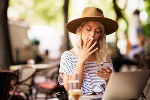 Mujer joven rubia con sombrero fuma un cigarrillo en una terraza y escribe mensajes de texto