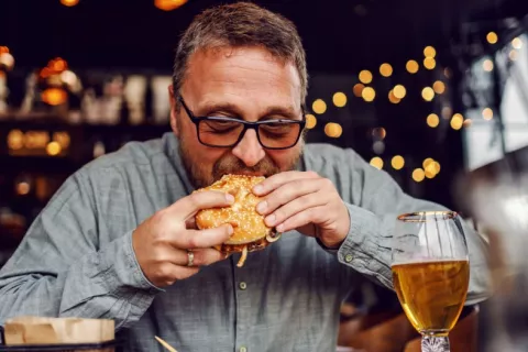 Hombre de mediana edad tomando una hamburguesa con patatas y cerveza Hombre de mediana edad devora una hamburguesa en un local de comida rápida
