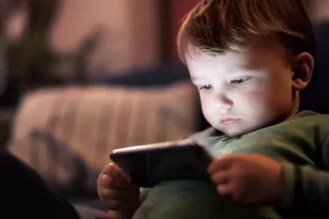 Niño pequeño en la cama a oscuras observa atentamente un smartphone Niño pequeño sentado en la cama mirando un móvil