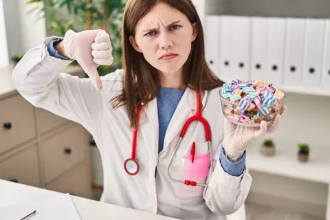 Joven doctora muestra caramelos y el pulgar hacia abajo Doctora sujeta un cuenco lleno de caramelos y hace un gesto de desacuerdo con el pulgar hacia abajo