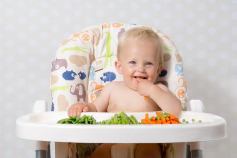 Un bebé en la trona come trozos de verduras cocidas que tiene delante
