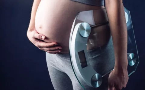 Mujer embarazada se toca el vientre con una mano y sujeta una báscula con la otra