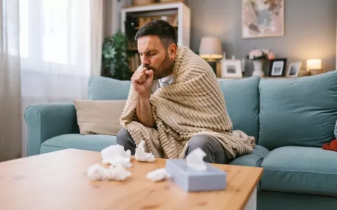 Hombre de mediana edad en su sofá arropado con una manta, con tos y pañuelos en la mesa