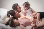 10 propuestas para introducir el slow parenting en tu familia Una pareja y sus dos hijas ríen tumbados en la cama