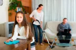 Cómo afectan a los niños los estereotipos de género Una mujer pasa la aspiradora y su hija limpia el polvo, mientras el padre consulta el ordenador
