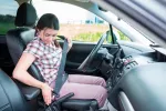 Cómo sentarte al volante para evitar lesiones y accidentes Mujer sentada en el coche colocándose el cinturón de seguridad para evitar lesiones y accidentes.