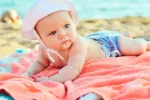 Los bebés menores de seis meses no deben estar al sol Bebé tumbado sobre una toalla en la playa con un gorrito en la cabeza