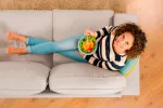 Dieta antiinflamatoria: para qué sirve y qué alimentos incluye Vista superior de una mujer en el sofá comiendo dieta antiinflamatoria