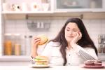 Una chica joven con sobrepeso mira una hamburguesa que tiene en la mano