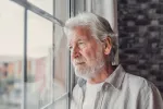 Hombre mayor con barba y canas mirando preocupado por la ventana