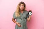 Edad biológica se acelera en las embarazadas Mujer rubia embarazada con cara de sorpresa y un reloj en la mano