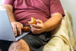 1.800 millones de personas pueden perder la salud por no hacer ejercicio Un adulto sentado en el sofá trabajando con el portátil y comiendo comida no saludable