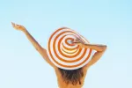 Cómo proteger tu cuero cabelludo y tu pelo de los rayos solares Chica de espaldas se sujeta el sombrero en la playa