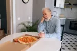 Vinculan las dietas inflamatorias con un 84% más riesgo de demencia Anciano sentado en la mesa frente una pizza con la mirada pensativa