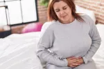 El analgésico no opioide Journvaux, primer fármaco de su clase aprobado por la FDA en 20 años Mujer joven con sobrepeso sentada en la cama se toca el abdomen con gesto de dolor