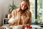 Cafeína, frutas, lácteos y fibra se asocian a menor riesgo de acúfenos Mujer joven vierte leche en sus cereales junto a taza de cafe y tazón de frutas