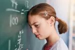 Niña en la escuela junto a la pizarra intenta resolver problema matemático Niña apoya la cabeza en la pizarra de la escuela con gesto de frustración