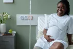 Joven embarazada en el hospital para un control prenatal Sonriente joven negra embarazada sentada sobre la cama del hospital