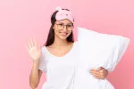 Chica sonriente preparada para acostarse temprano Chica con un antifaz en la frente y sujetando una almohada se despide sonriente antes de irse a la cama